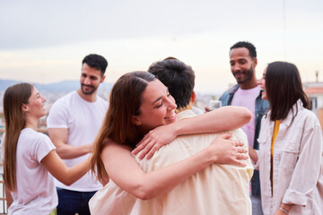Two people embraced outdoors during a meeting. Female and male friends hugging surrounded by a group of people gathered outside in a rooftop. Greeting at a party.