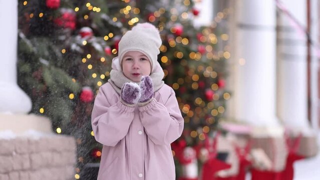Happy Caucasian child girl blows snow from her hands. A child is having fun outdoors in winter against the backdrop of a Christmas tree and garland lights.