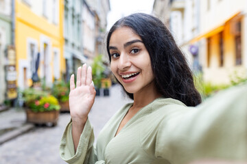 Close-up portrait of an Indian young woman traveling, walking on a city street, filming herself on the phone, talking on a video call, writing a blog, waving and saying hello to the camera © Tetiana