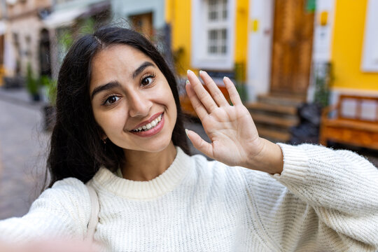 A beautiful young Indian woman in a white sweater is standing on a city street and talking on a video call on a telephone, waving and saying hello