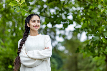 A happy and smiling Indian woman in a white sweater and a braided hairstyle stands in the park with her arms crossed on her chest and looks confidently to the side
