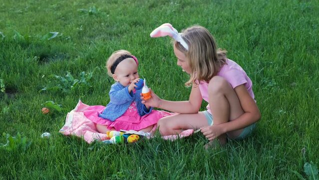 Baby girl with older sister playing with colorful eggs on the grass outdoors. Easter hunt concept