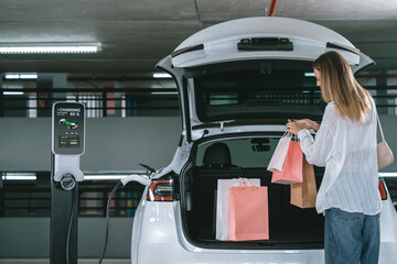 Young woman travel with EV electric car to shopping center parking lot charging in downtown city...