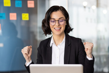 Successful female in formal suit doing yes gesture with clenched fist while looking at laptop...