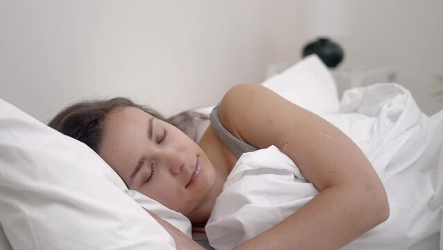 Serenely asleep, a young lady rests under a white blanket on her comfortable orthopedic mattress in a well-lit bedroom.