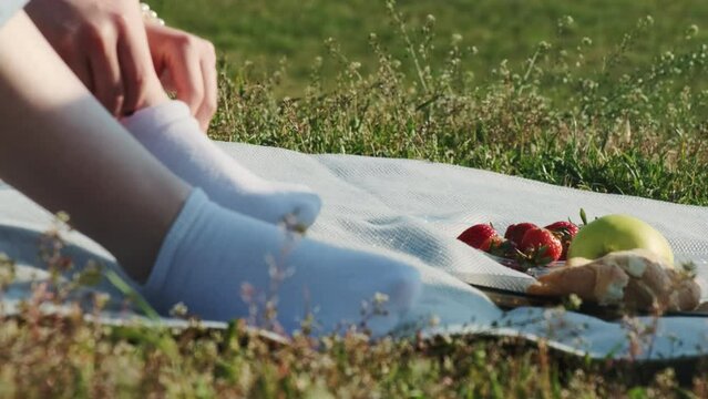 Woman in jeans puts on sock sitting on blanket during picnic. Summer picnic with juicy fruits and fresh pastry on sunny day