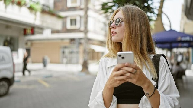 A young attractive woman with blonde hair and sunglasses uses a smartphone on a sunny city street.