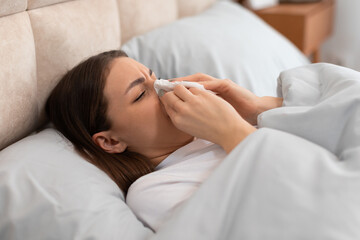 Young woman with cold, blowing her nose, lying in bed - obrazy, fototapety, plakaty