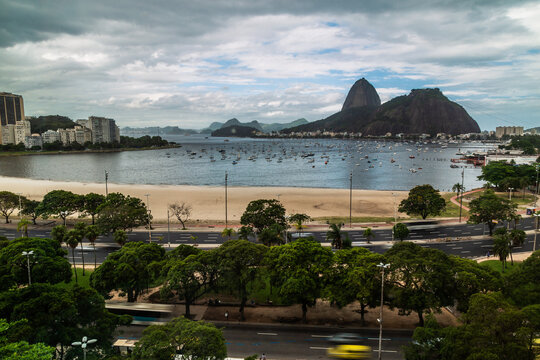 Vista panor&acirc;mica da Marina da Gl&oacute;ria, no Rio de Janeiro, com o P&atilde;o de A&ccedil;&uacute;car ao fundo, &aacute;rvores ao redor e c&eacute;u parcialmente nublado, ideal para turismo e lazer