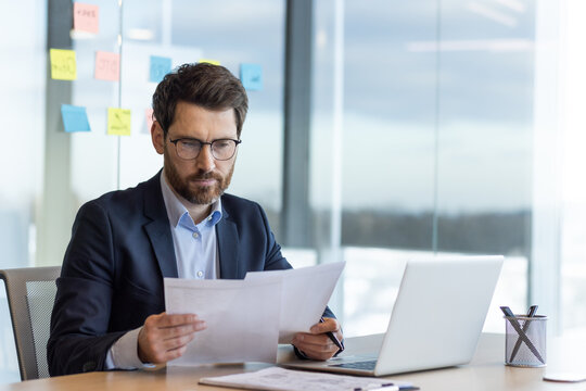 Serious thinking businessman behind paper work, mature experienced boss working with reports and financial documents, financier inside office with laptop, reading contract, thoughtfully checking.