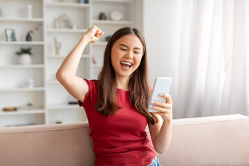 Euphoric young asian woman cheering success with smartphone at home