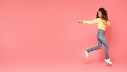 Excited black woman jumps, pointing aside at copy space on pink backdrop, panorama