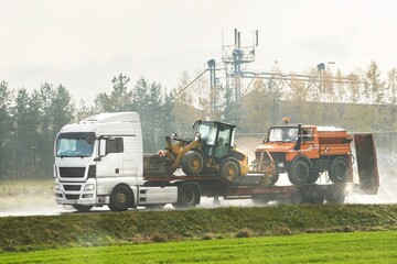 A white truck hauls heavy construction equipment on a trailer. Loaders and bulldozers are transported by a white truck ready for road construction.