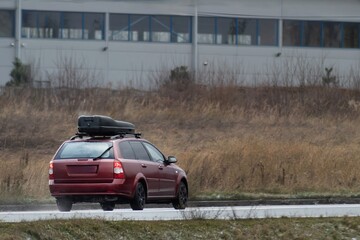 A modern car with a roof box travels down a road surrounded by grasslands under an overcast sky.