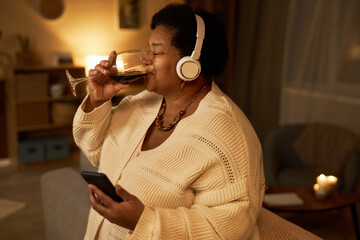 Side view portrait of adult Black woman drinking wine at home and listening to music with headphones