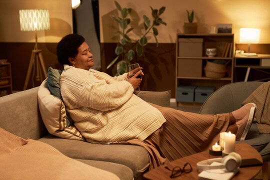 Full length side view of senior Black woman relaxing on sofa at home with feet up and holding glass of wine, copy space