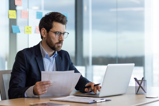 Serious and focused financier accountant on paper work inside office, mature man using calculator and laptop for calculating reports and summarizing accounts, businessman at work in suit.