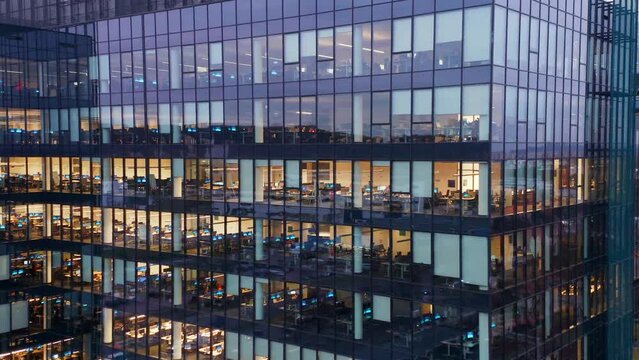 Cinematic drone shot office windows in a glass skyscraper at night close-up. People working in office building in the night, building lights. Workers at office building at night, view trough a window