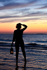 A dark silhouette of a girl with a lantern in her hand against the background of the sea and a beautiful sunset