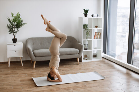Brunette staying in headstand pose at bright commodious living room. Fit and slender lady doing yoga exercises in beige tight leggings and top in front of french windows.