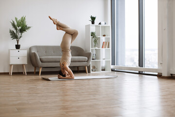 Brunette staying in headstand pose at bright commodious living room. Fit and slender lady doing...