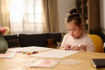 Adorable child with mental disability drawing with crayons on paper while sitting by table in front...