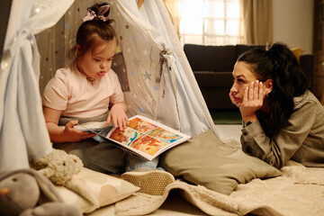 Young woman looking at her adorable little daughter with Down syndrome sitting in tent and looking at pictures in book of comics