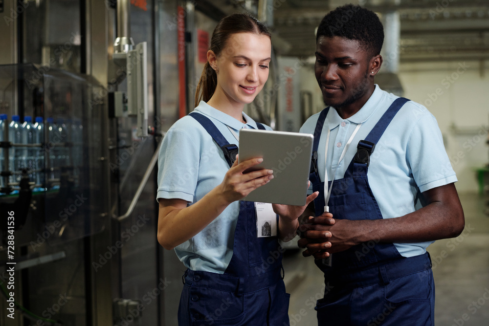 Poster two young confident quality control inspectors in workwear looking at screen of tablet held by woman