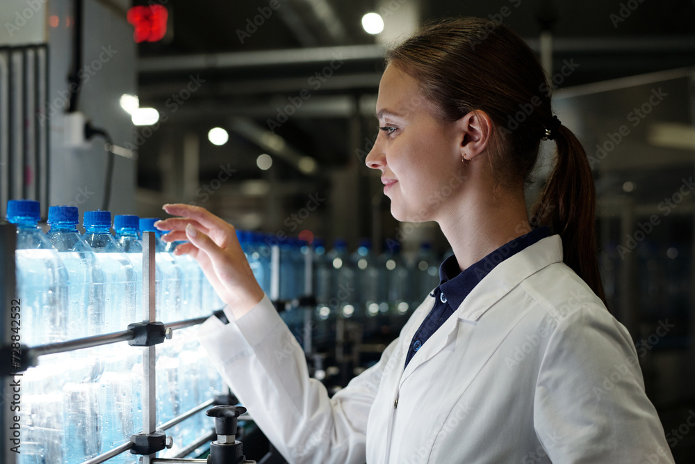 Aufkleber young female quality inspector in labcoat standing in front of assembly line with capped bottled dri