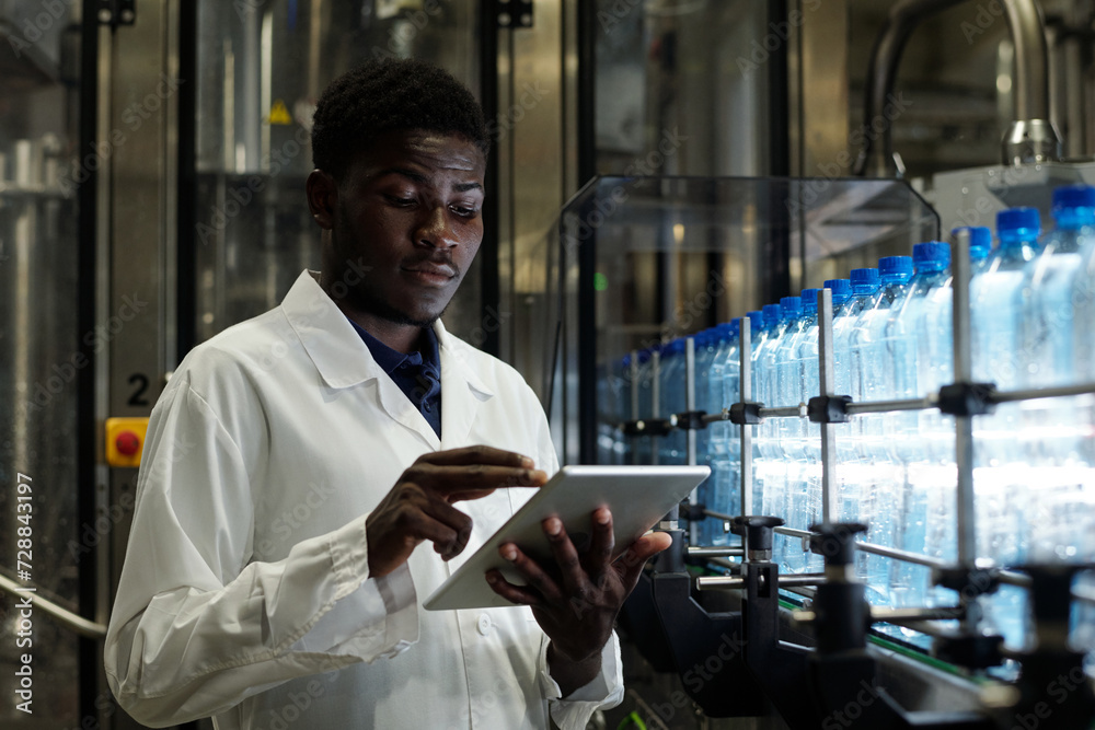 Aufkleber Young male inspector of factory in labcoat standing in front of conveyor belt with capped bottled drinks and scrolling in tablet