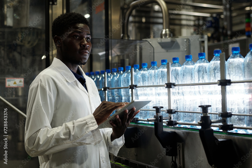 Poster quality control inspector with tablet standing in front of conveyor belt with capped bottles of mine