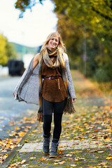 A young, blonde woman in a knitted look walks towards the camera along a sidewalk covered with...