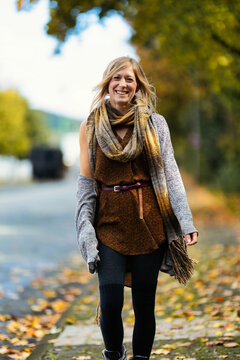 A young, blonde woman in a knitted look walks towards the camera along a sidewalk covered with autumn leaves.
