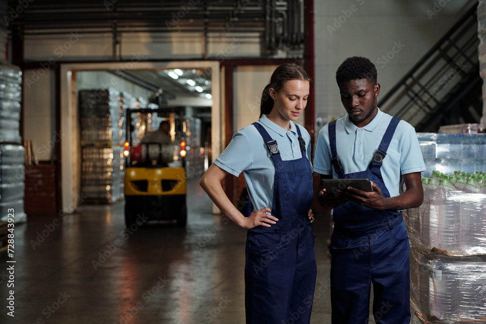Poster Two young intercultural engineers of quality control looking at screen of tablet while checking or discussing information in warehouse