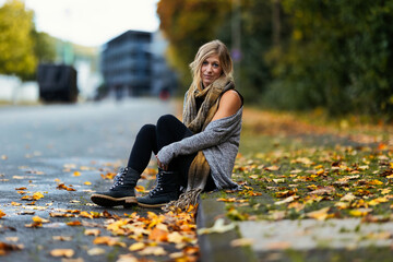 A young blonde woman in a knitted earth-colored look with black leggings and blue boots sits on a...