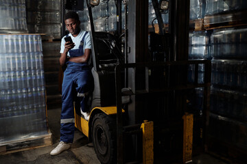 Young factory engineer in coveralls sitting by door of forklift after work and texting in his smartphone against stacks of bottled water