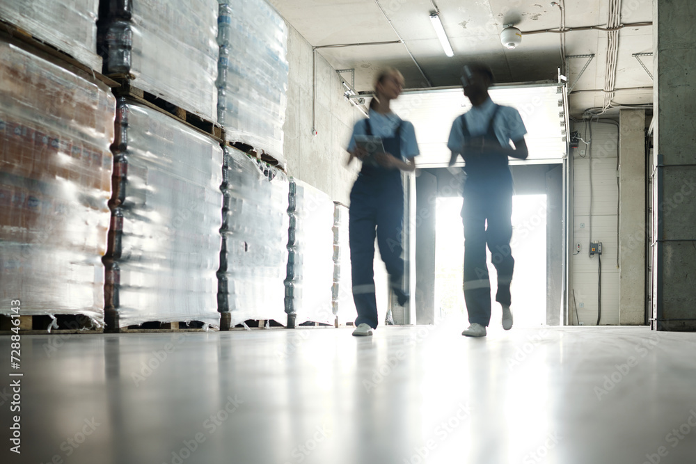 Poster blurry outlines of two engineers of bottled water factory moving along stacks of huge packages in sp
