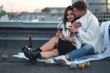 Surprise date on rooftop with urban cityscape and skyscrapers on background. Happy young loving couple drinking wine having romantic candlelit dinner celebrating anniversary or Valentines Day