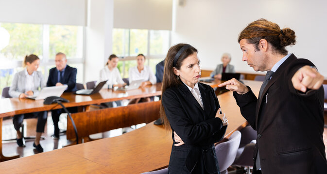 Disgruntled annoyed middle aged white male boss pointing an incompetent upset middle aged white brunette female employee to the door while other coworkers sitting and watching them during team meeting