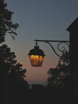 Street lamp lit on the street with trees and sunset in the background.