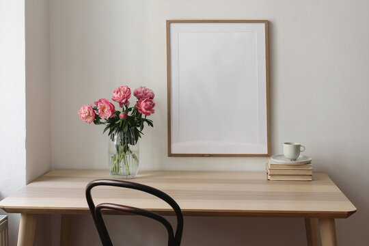 Elegant interior still life. Blank vertical picture frame mockup. Vase with pink peonies flowers. Cup of tea, coffee on books. Wooden table, desk. Romantic home breakfast. White wall background.
