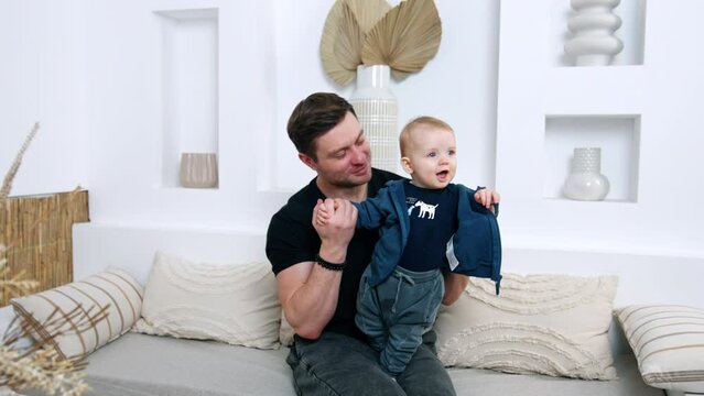 Man in black t-shirt sits on the sofa holding his son. Little kid smiles to camera, dad kisses and hugs his adorable baby.
