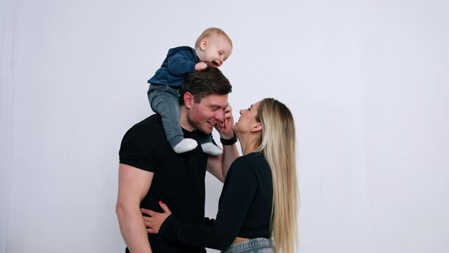Cute little infant boy sits on the shoulders of his father. Mom tries to reach her baby playing and kissing him. White backdrop.