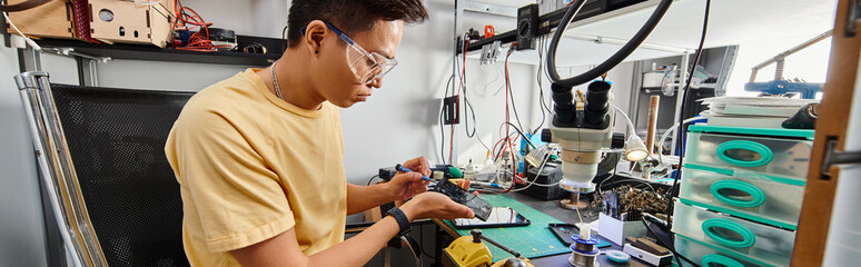 skilled asian technician in goggles working with electronic devices in repair workshop, banner