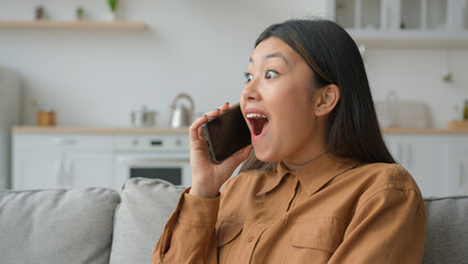 Excited Asian woman businesswoman talk phone in kitchen listen good news feel shock surprised...