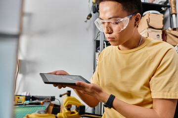 attentive asian repairman in goggles holding digital tablet with broken touchscreen in workshop