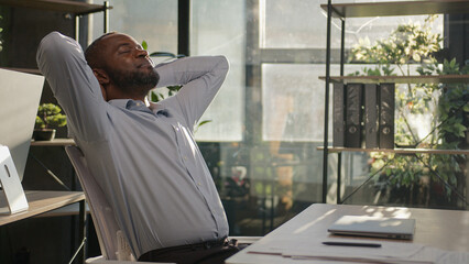 Satisfied businessman African American mature middle-aged 45s man finish computer project close laptop work day off lean back on chair put hands behind head take break relaxing in sunshine warm office - obrazy, fototapety, plakaty