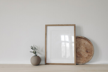 Blank vertical picture frame mockup, poster display. Ball vase with olive tree branch. Round wooden tray on table, desk. Minimal rustic home. Scandinavian interior. White wall background. - obrazy, fototapety, plakaty