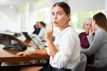 Strict young white business woman making shush gesture with index finger over her mouth demanding silence while sitting at corporate team meeting 