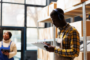 African american young shipment manager wearing headphones taking notes in warehouse. Storehouse...